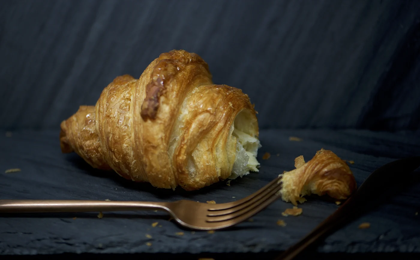 A golden L’Artisane croissant on a dark tabletop beside a fork and a small piece of pastry