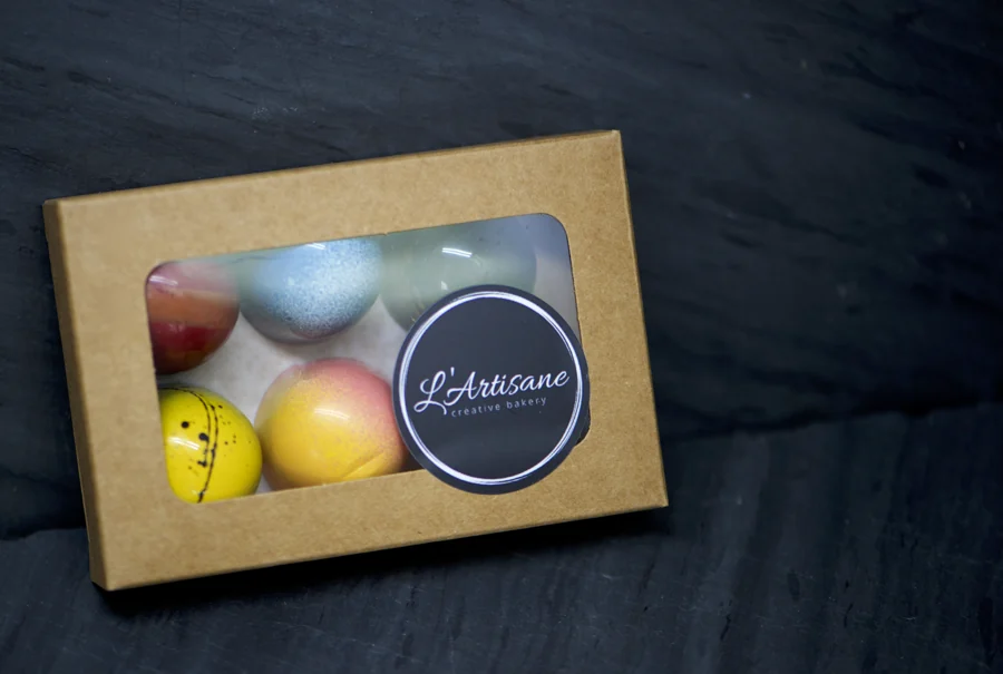 A box of colorful L’Artisane macarons bearing the original circular bakery logo